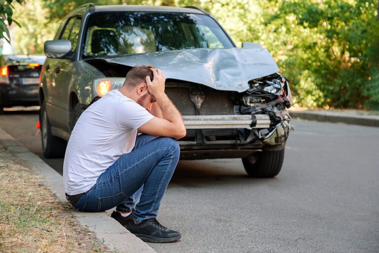 A man is seated on the ground beside a car with visible damage, considering important post-accident procedures for drivers.